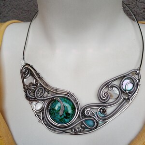 May include: A handcrafted silver wire necklace with a large turquoise stone and smaller turquoise and pearl accents. The necklace has an artistic design with intricate wirework and a curved shape.