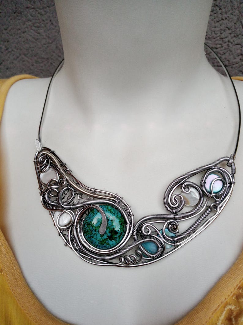 May include: A handcrafted silver wire necklace with a large turquoise stone and smaller turquoise and pearl accents. The necklace has an artistic design with intricate wirework and a curved shape.