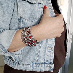 May include: A silver-toned wire-wrapped bracelet with red beads. The bracelet features an intricate design with a central circular element and smaller beads. The jewelry is worn on a person's wrist, against a denim jacket.