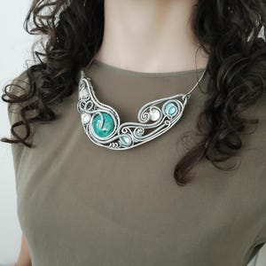 May include: A silver wire necklace with a central turquoise stone and smaller turquoise and white beads. The necklace has a flowing, organic design. The necklace is a statement piece.