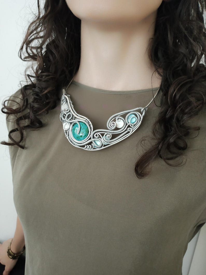 May include: A silver wire necklace with a central turquoise stone and smaller turquoise and white beads. The necklace has a flowing, organic design. The necklace is a statement piece.