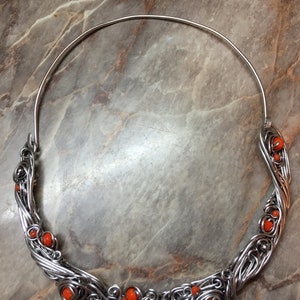 May include: A silver wire necklace with orange beads. The necklace is made of wire that is twisted and woven together to create a unique design. The beads are small and round, and they are evenly spaced along the necklace.