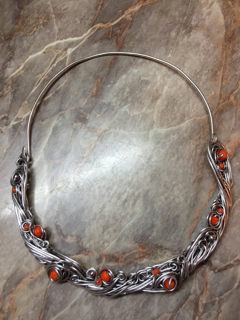 May include: A silver wire necklace with orange beads. The necklace is made of wire that is twisted and woven together to create a unique design. The beads are small and round, and they are evenly spaced along the necklace.