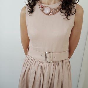 May include: A sleeveless beige dress with a pleated skirt and a matching belt featuring a silver-toned rectangular buckle. The dress is paired with a copper wire-wrapped statement necklace containing a large round white stone and small red accents.