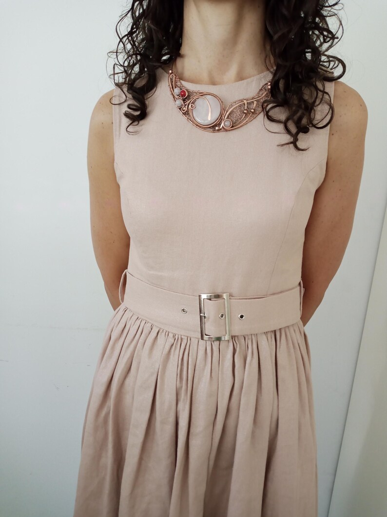May include: A sleeveless beige dress with a pleated skirt and a matching belt featuring a silver-toned rectangular buckle. The dress is paired with a copper wire-wrapped statement necklace containing a large round white stone and small red accents.