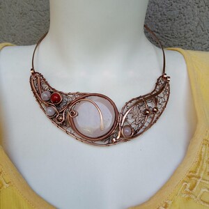 May include: A copper wire-wrapped collar necklace featuring a large, central, circular pale pink stone. The design includes intricate wire mesh detailing, two small pale pink beads, and one vibrant red bead, all set within a structured copper frame.