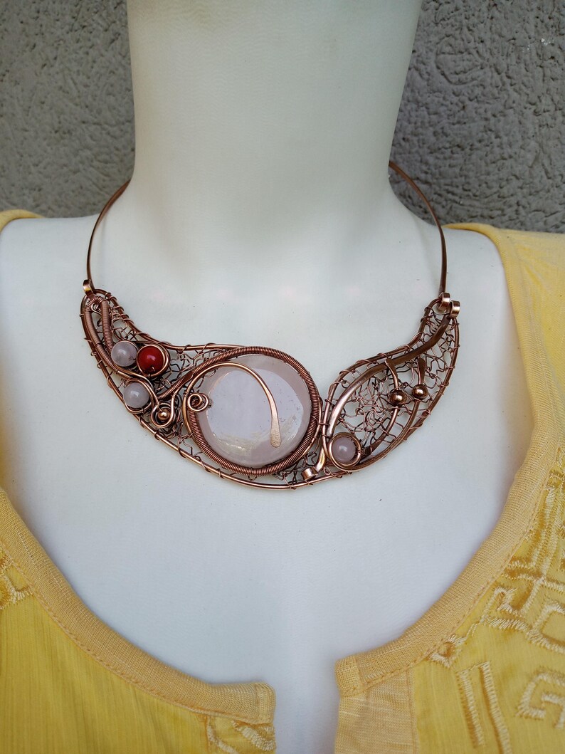 May include: A copper wire-wrapped collar necklace featuring a large, central, circular pale pink stone. The design includes intricate wire mesh detailing, two small pale pink beads, and one vibrant red bead, all set within a structured copper frame.