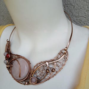 May include: A copper wire-wrapped collar necklace featuring a large, round, pale pink stone centerpiece. The design includes intricate woven copper wire patterns, small spherical copper beads, and two additional small, round, pale pink and red stones.