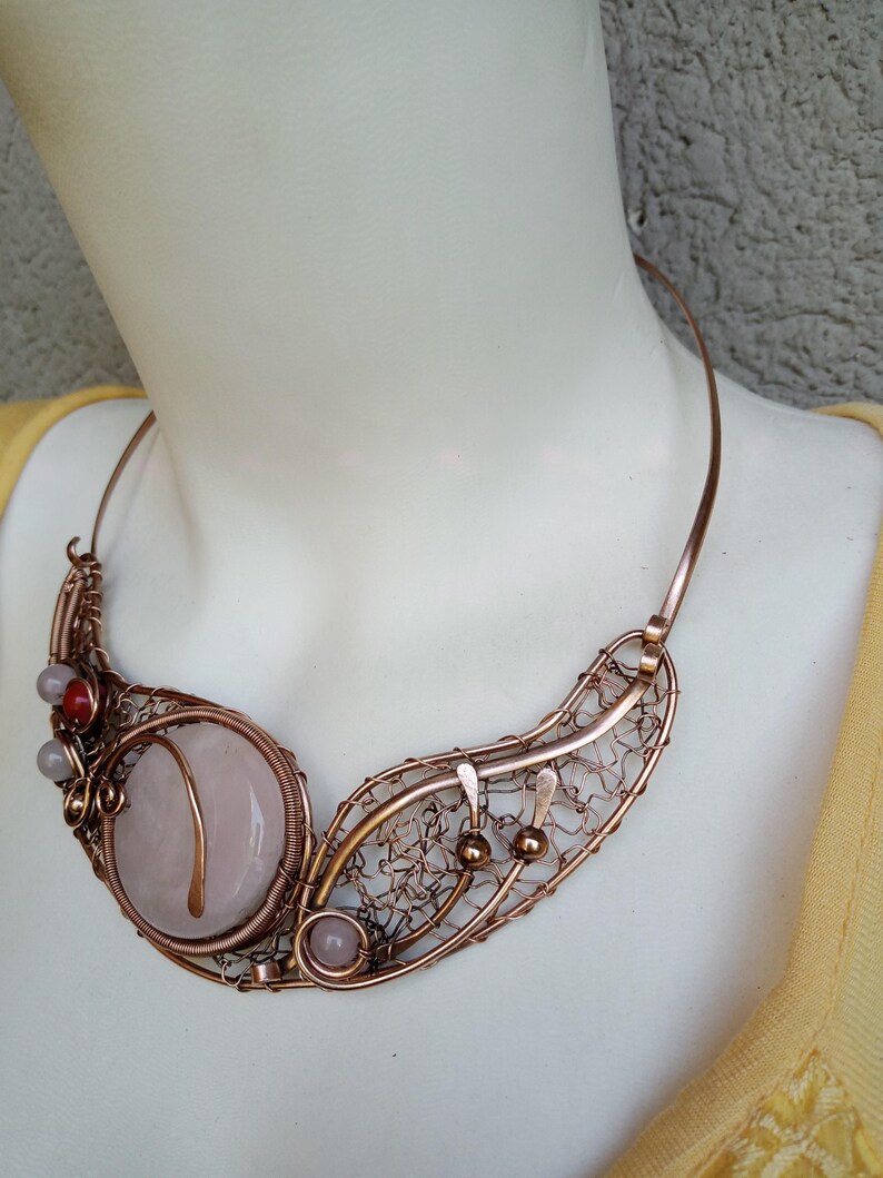 May include: A copper wire-wrapped collar necklace featuring a large, round, pale pink stone centerpiece. The design includes intricate woven copper wire patterns, small spherical copper beads, and two additional small, round, pale pink and red stones.
