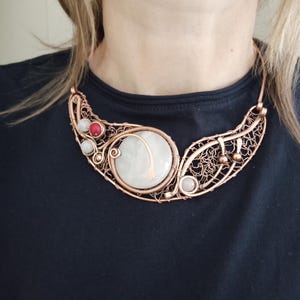 May include: A copper wire-wrapped collar necklace featuring a large central circular white stone, accented with smaller round white and red beads. The metalwork consists of intricate, swirling wire patterns that form a crescent-shaped frame.