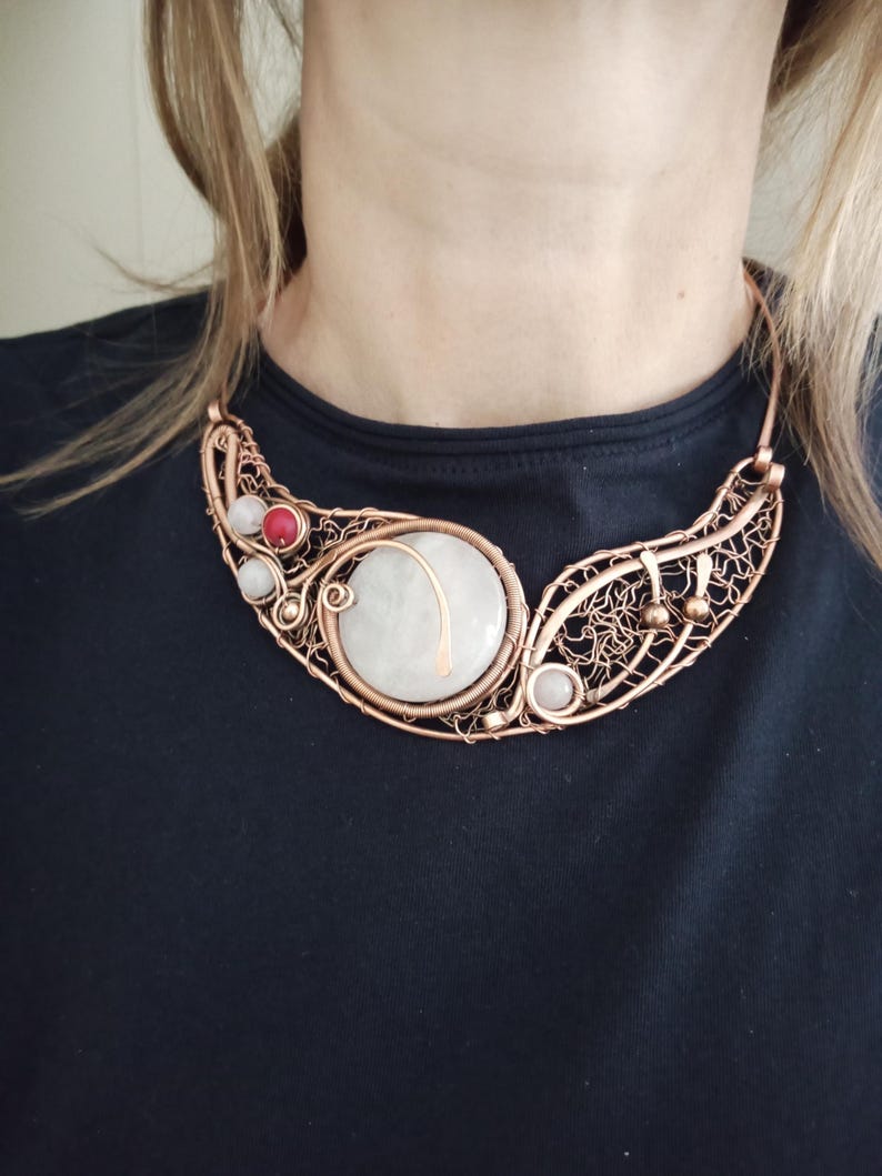 May include: A copper wire-wrapped collar necklace featuring a large central circular white stone, accented with smaller round white and red beads. The metalwork consists of intricate, swirling wire patterns that form a crescent-shaped frame.