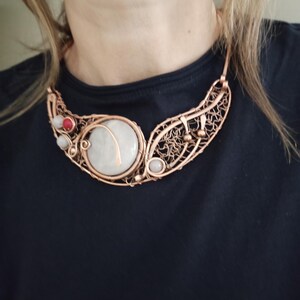 May include: A copper wire-wrapped collar necklace featuring a large, round, off-white stone centerpiece, accented with smaller red and white beads. The design incorporates intricate, woven copper wire patterns and metal spheres along the curved frame.
