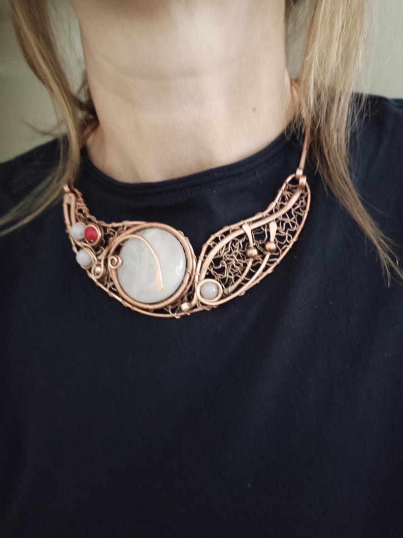 May include: A copper wire-wrapped collar necklace featuring a large, round, off-white stone centerpiece, accented with smaller red and white beads. The design incorporates intricate, woven copper wire patterns and metal spheres along the curved frame.