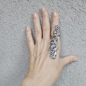 May include: Two silver rings with detailed, swirling patterns. One ring extends along the finger, the other is a smaller band. The rings are shown on a hand against a textured grey background. The rings appear to be made of metal.