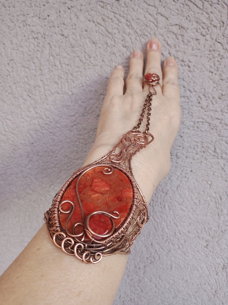 Pu&ograve; includere: Un braccialetto in filo di rame con una grande pietra rossa di forma ovale. Il braccialetto &egrave; collegato a un anello con una pietra rossa pi&ugrave; piccola. Il braccialetto &egrave; progettato con un intricato lavoro a filo e ha un aspetto delicato e femminile.