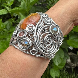 Könnte beinhalten: Ein breites Armband aus silberfarbenem Draht mit komplexen Wirbelmustern und Schlaufen. Das Design enthält einen großen, glatten, orangefarbenen Stein sowie mehrere kleinere, facettierte, irisierende Glasperlen, die in das Drahtgeflecht eingearbeitet sind.