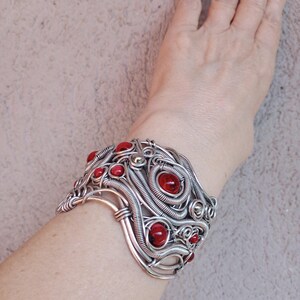 May include: A silver-toned wire-wrapped cuff bracelet with multiple red, round gemstones. The bracelet has an intricate, swirling design and is worn on a wrist. The gemstones are evenly spaced throughout the design.