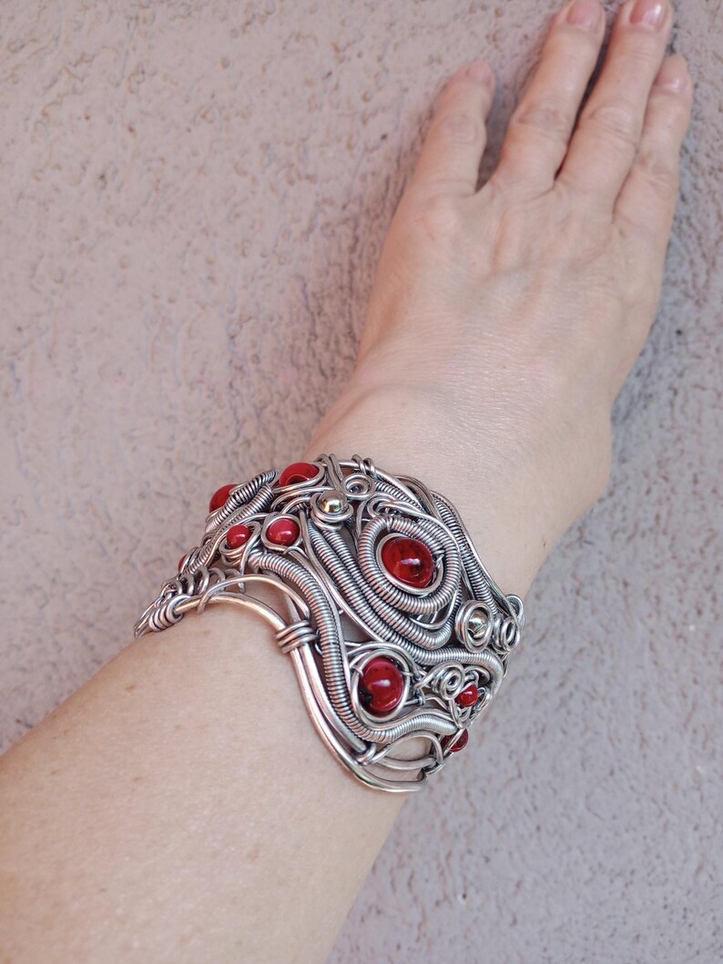 May include: A silver-toned wire-wrapped cuff bracelet with multiple red, round gemstones. The bracelet has an intricate, swirling design and is worn on a wrist. The gemstones are evenly spaced throughout the design.