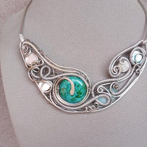 May include: A handcrafted silver wire necklace with a central turquoise stone. The necklace has an intricate, openwork design with smaller white and blue stones. The necklace is displayed against a neutral background.