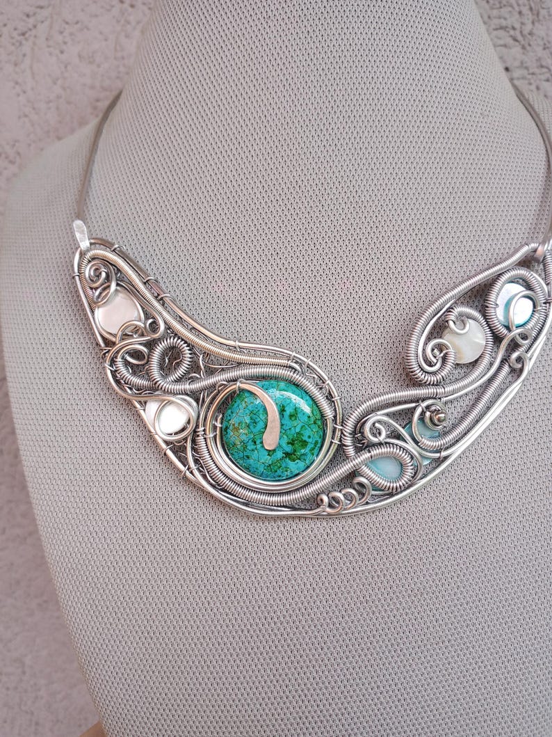 May include: A handcrafted silver wire necklace with a central turquoise stone. The necklace has an intricate, openwork design with smaller white and blue stones. The necklace is displayed against a neutral background.