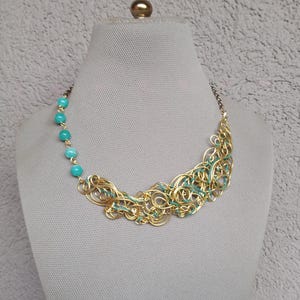 May include: A gold-coloured wire necklace with turquoise beads and teal accents. The necklace is displayed on a mannequin. The design features intricate wirework and a row of turquoise beads on one side, creating a unique and artistic jewellery piece.
