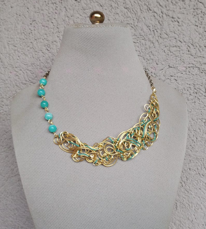May include: A gold-coloured wire necklace with turquoise beads and teal accents. The necklace is displayed on a mannequin. The design features intricate wirework and a row of turquoise beads on one side, creating a unique and artistic jewellery piece.
