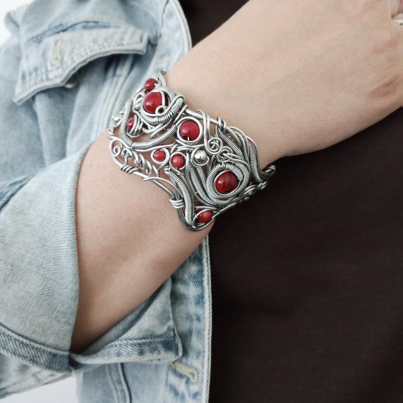 May include: A silver-toned cuff bracelet with intricate wirework and embedded red stones. The bracelet has a wide design, with the red stones varying in size and shape. The bracelet is worn on a person's arm, against a backdrop of a denim jacket and a brown shirt.