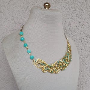May include: A gold-coloured necklace with turquoise beads and a woven design. The necklace features a series of turquoise beads on one side and a cluster of gold-coloured wire with teal accents on the other. The necklace is displayed on a mannequin.