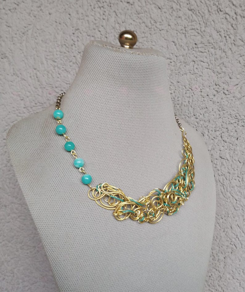 May include: A gold-coloured necklace with turquoise beads and a woven design. The necklace features a series of turquoise beads on one side and a cluster of gold-coloured wire with teal accents on the other. The necklace is displayed on a mannequin.