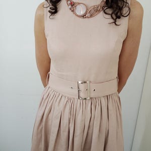May include: A sleeveless beige dress with a gathered waist and a matching belt featuring a silver-toned rectangular buckle. The dress is paired with a copper-toned wire-wrapped statement necklace containing a large central white stone and small red accents.