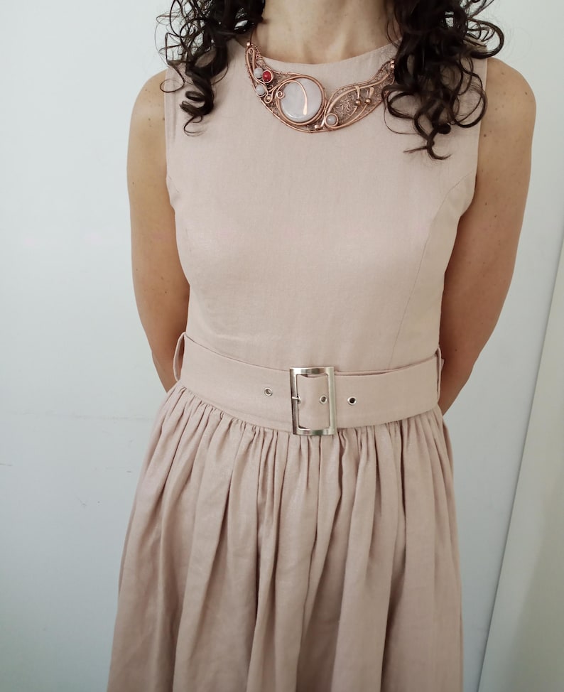 May include: A sleeveless beige dress with a gathered waist and a matching belt featuring a silver-toned rectangular buckle. The dress is paired with a copper-toned wire-wrapped statement necklace containing a large central white stone and small red accents.