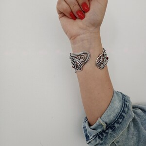 May include: A silver-toned cuff bracelet with an open design. The bracelet features intricate, swirling patterns and small red accents. The cuff is worn on a wrist, with a denim jacket sleeve visible in the background.