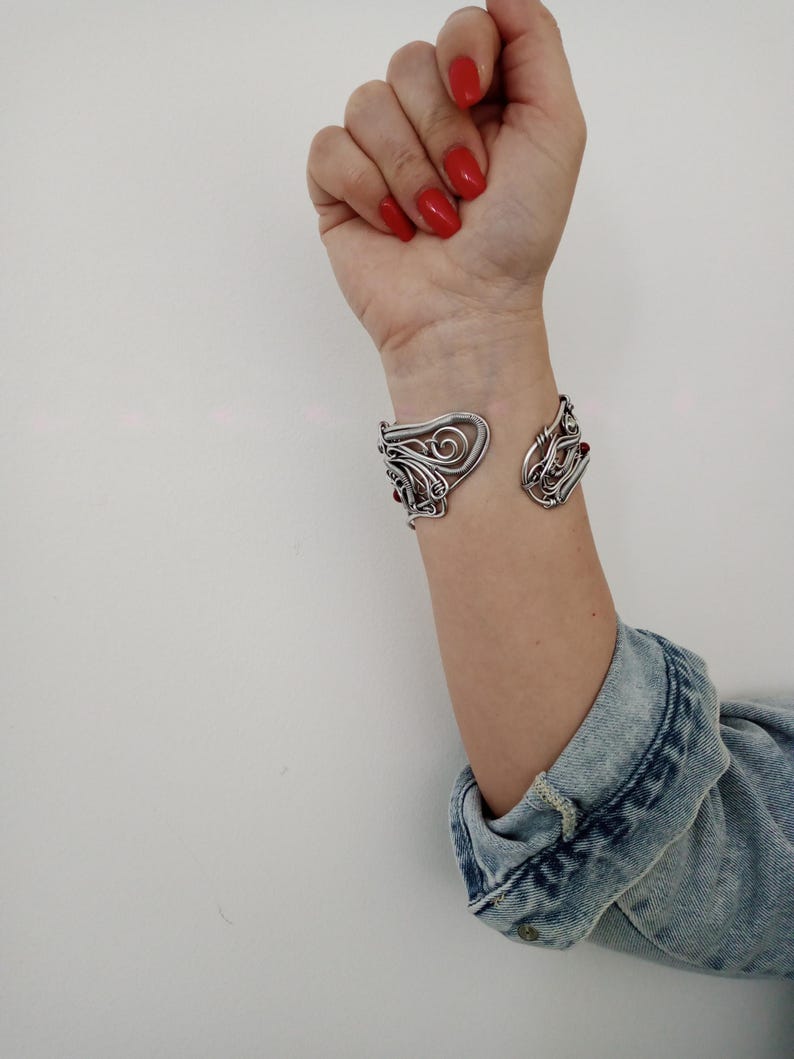 May include: A silver-toned cuff bracelet with an open design. The bracelet features intricate, swirling patterns and small red accents. The cuff is worn on a wrist, with a denim jacket sleeve visible in the background.