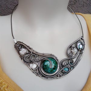 May include: A handcrafted silver wire necklace with a large, round, green stone centre. The necklace features intricate wirework with smaller stones and shell accents. The necklace is displayed on a mannequin.