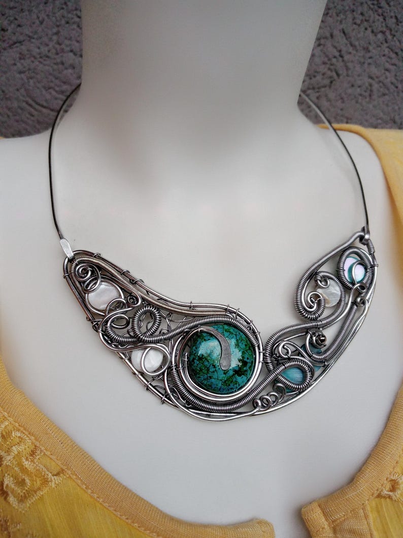 May include: A handcrafted silver wire necklace with a large, round, green stone centre. The necklace features intricate wirework with smaller stones and shell accents. The necklace is displayed on a mannequin.