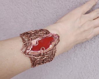 May include: A copper wire cuff bracelet featuring a large, polished red and white agate stone. The stone is the focal point, set within a woven copper wire frame. The bracelet is designed to be worn on the wrist.