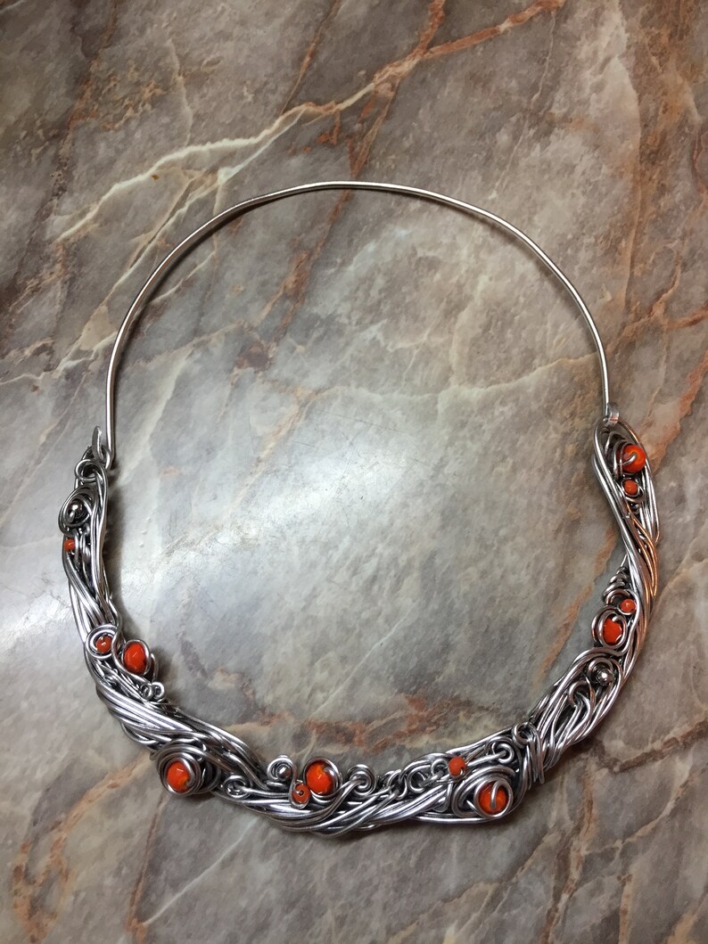 May include: A silver wire necklace with orange beads. The necklace is made of wire that is twisted and woven together to create a unique design. The beads are small and round, and they are evenly spaced along the necklace.
