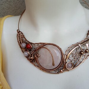 May include: A wire-wrapped copper necklace featuring a large, round, pale pink stone centerpiece. The design incorporates intricate copper wire filigree, small light pink beads, a single red bead, and metallic accents, forming a wide, curved collar.