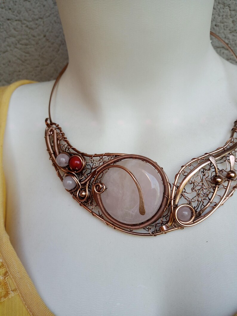 May include: A wire-wrapped copper necklace featuring a large, round, pale pink stone centerpiece. The design incorporates intricate copper wire filigree, small light pink beads, a single red bead, and metallic accents, forming a wide, curved collar.