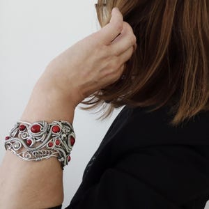 May include: A silver-toned wire-wrapped bracelet with multiple red gemstone accents. The bracelet features an intricate design with swirling patterns and is worn on a person's arm. The background is a plain white wall.