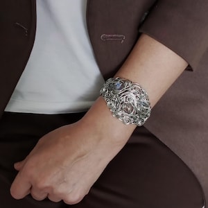 May include: A silver-coloured wire-wrapped cuff bracelet with three large, iridescent gemstones. The bracelet has intricate swirling wire designs. The person is wearing a brown blazer and a white shirt.