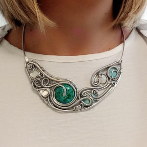 May include: A silver wire-wrapped necklace with a large, round, green stone at the centre. The necklace has a flowing design with smaller turquoise and white stones. The necklace is worn around the neck.