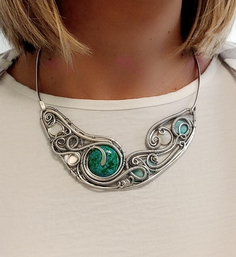 May include: A silver wire-wrapped necklace with a large, round, green stone at the centre. The necklace has a flowing design with smaller turquoise and white stones. The necklace is worn around the neck.