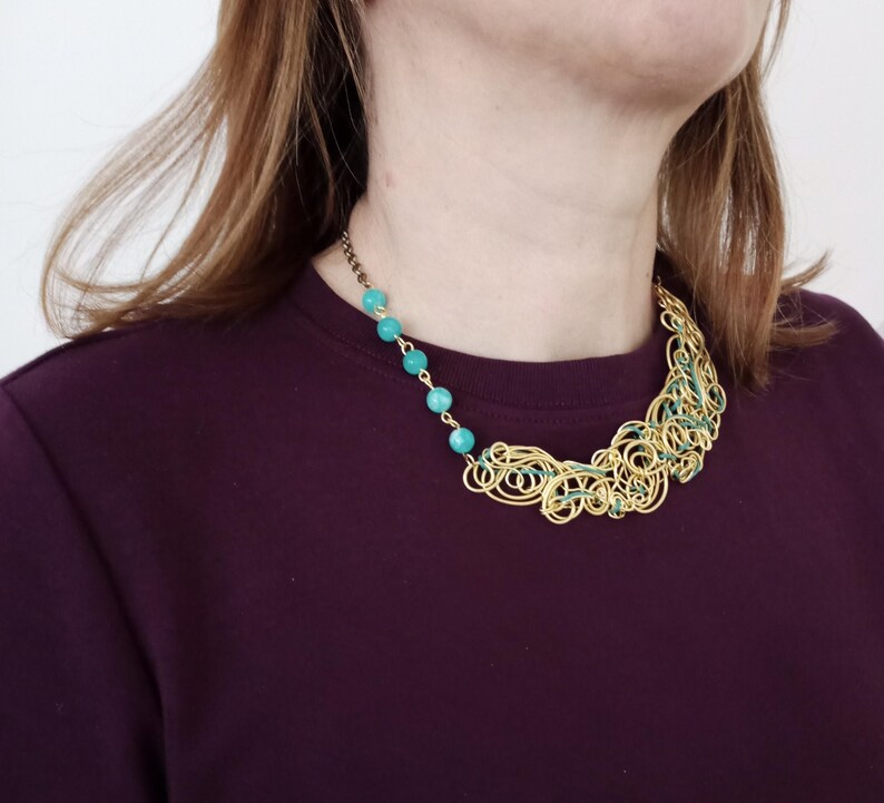 May include: A gold-coloured wire necklace featuring turquoise beads and green detailing. The necklace has an abstract design with loops and curves. Turquoise beads are strung along a chain on one side, creating a striking contrast.