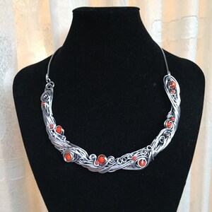 May include: A silver wire necklace with orange beads. The necklace is in the shape of a wave and has a delicate, intricate design.