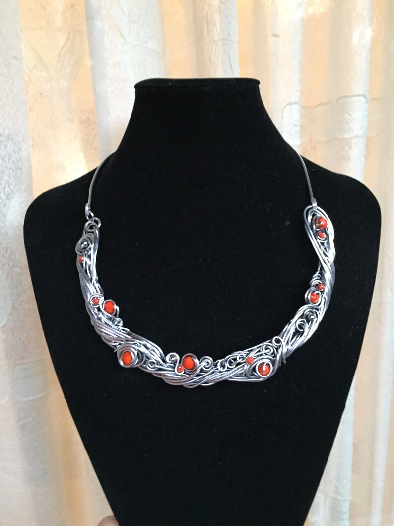 May include: A silver wire necklace with orange beads. The necklace is in the shape of a wave and has a delicate, intricate design.