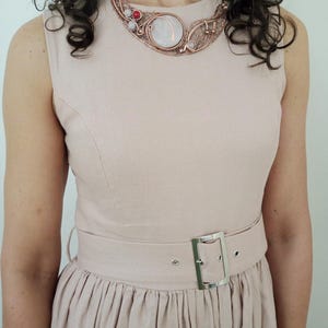 May include: A sleeveless light pink dress with a round neckline and a matching belt featuring a silver rectangular buckle. The dress is paired with a copper-toned wire-wrapped statement necklace containing a large central stone and small red accents.