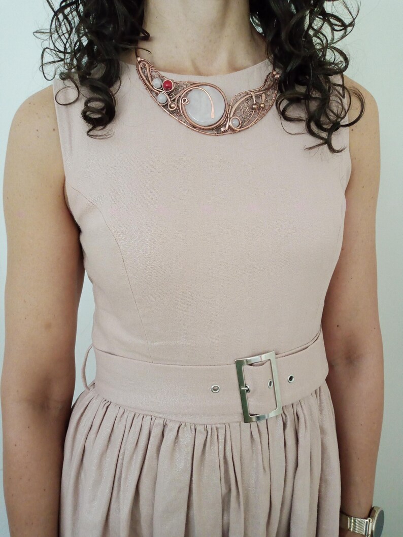 May include: A sleeveless light pink dress with a round neckline and a matching belt featuring a silver rectangular buckle. The dress is paired with a copper-toned wire-wrapped statement necklace containing a large central stone and small red accents.