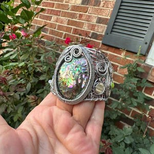 May include: A wire-wrapped cuff bracelet featuring a large, oval-shaped iridescent abalone shell centrepiece. The silver-toned metal wire is intricately coiled and woven into a complex, open-work lattice design surrounding the central shell.