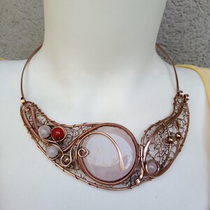 May include: A wire-wrapped copper necklace featuring a large, round, pale pink stone centerpiece. The design includes intricate copper wire filigree, smaller light pink beads, and a single vibrant red bead, all set on a rigid copper collar.